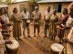Groupe mixte de musiciens réunionnais en cercle, avec un roulèr au premier plan, des kayambs et des chanteurs vêtus sobrement dans une lumière naturelle chaleureuse.