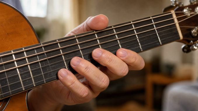 Gros plan sur la main gauche d’un homme formant proprement un accord de si mineur sur une guitare acoustique.