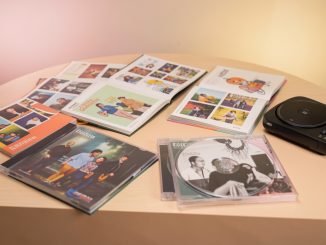 Table de collectionneur présentant quatre albums de musique physiques ouverts, des photocards colorées, des livrets photo et un lecteur CD sous une lumière douce de studio.
