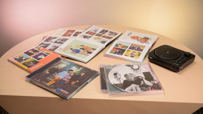 Table de collectionneur présentant quatre albums de musique physiques ouverts, des photocards colorées, des livrets photo et un lecteur CD sous une lumière douce de studio.